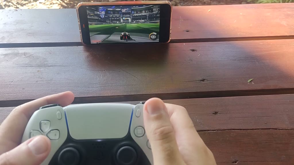 Un telefono appoggiato su un tavolo da picnic con Rocket League in streaming tramite LinkRaze, e un controller wireless tenuto con entrambe le mani.